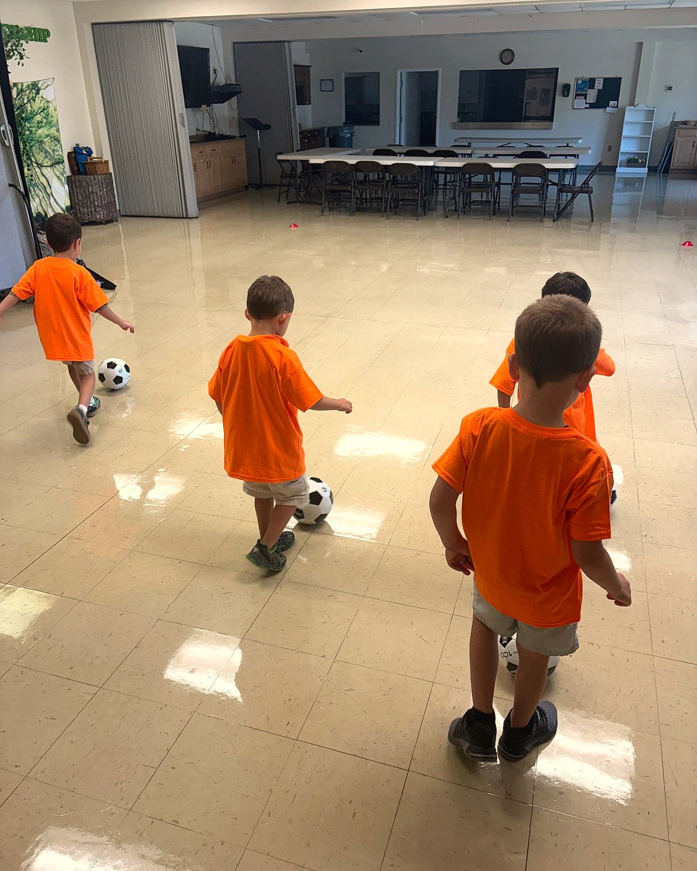 Cubs in matching orange shirts kicking soccer balls across a daycare hall during class